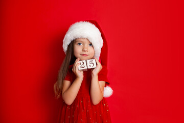 Merry Christmas and Happy Holidays. A funny girl in Santa's hat holds cubes with the numbers 25. Red background, space for text.