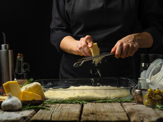 The chef grates cheese onto the dough prepared for baking. Ingredients for making pie, focaccia, pizza. Wooden texture. Country style. Cookbook, culinary blog, restaurant.