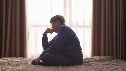 Fototapeta premium Depressed lonely old elderly Asian woman, people sitting on bed with windows in bedroom at home in early morning. Quarantine lifestyle. Unhappy life.