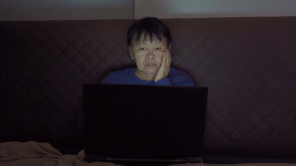 Depressed elderly old Asian woman, people using a notebook laptop computer on social media internet on bed in bedroom at home. Lifestyle on late night in technology device concept. Insomnia