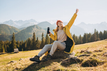 Traveler girl drinking tea from thermos cup over nature background. Mountain peaks. Freedom, happiness, travel and vacations concept, outdoor activities, woman wearing red hat