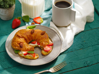 Tasty breakfast. Buttered French croissant, sprinkled with almonds and fresh strawberries on top, a cup of black coffee, milk, decor. Nicely served table for one person. Home comfort.