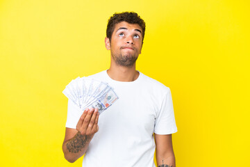 Young brazilian man taking a lot of money isolated on yellow background and looking up
