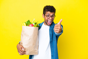 Young Brazilian man holding a grocery shopping bag isolated on yellow background with thumbs up...