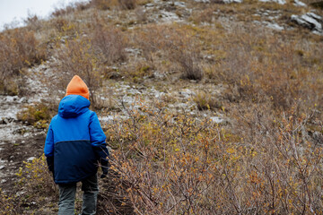 Fototapeta na wymiar The child goes camping. A boy in a jacket climbs uphill. View from the back of a child walking on the road to the top. Children's tourism in autumn in cloudy weather.