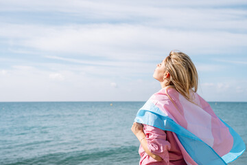 Woman standing on seashore on windy day holding transgender flag