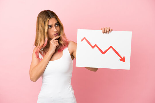 Young woman over isolated pink background holding a sign with a decreasing statistics arrow symbol and thinking