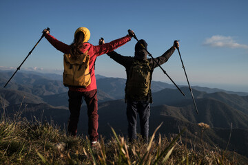 Fototapeta na wymiar Man and woman travelers in red jacket with yellow backpack are standing with their backs turned and looking at mountains with arms outstretched sticks to sides. Travel in mountains with loved one.