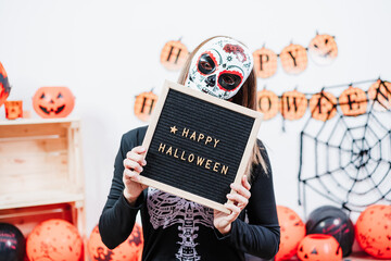 woman wearing mexican face mask during halloween celebration. woman holding letter board wearing skeleton costume and lying on the floor pretending to be dead. Halloween party concept