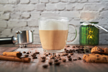A cappuccino surrounded by coffee beans, cinnamon sticks and cookies. Shot from the front on a wooden table. Portafilter can be seen in the background.