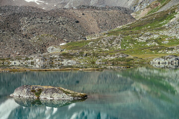 Rock in turquoise mountain lake. Snowy mountain reflected in azure clear water of glacial lake. Beautiful sunny background with snow-white glacier reflection in green water surface of mountain lake.