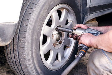 A mechanic at a service station replaces a car wheel, a worker's hands in black fuel oil and a tool for unscrewing bolts close-up near the wheel. Tire service, workflow