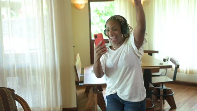 Mujer afroamericana bailando mientras escucha m&uacute;sica con el celular conectado a los aud&iacute;fonos en su casa