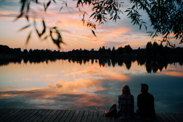 Romantic holiday. Young loving couple sitting together on lake bank enjoying beautiful sunset.