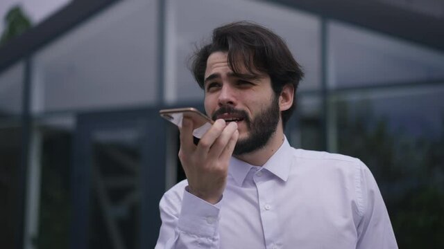 Concentrated handsome bearded man recording voice mail on smartphone standing on office terrace outdoors. Portrait of confident serious Caucasian employee sending message in app online