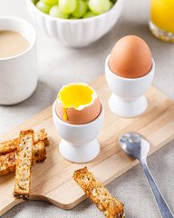 Two soft-boiled eggs in egg cups served on wooden cutting board with toasted bread for breakfast. Food background.