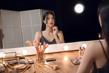 Beautiful young woman applying lipstick with brush in dressing room at night