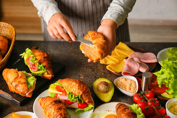Woman preparing tasty croissant sandwich at kitchen table