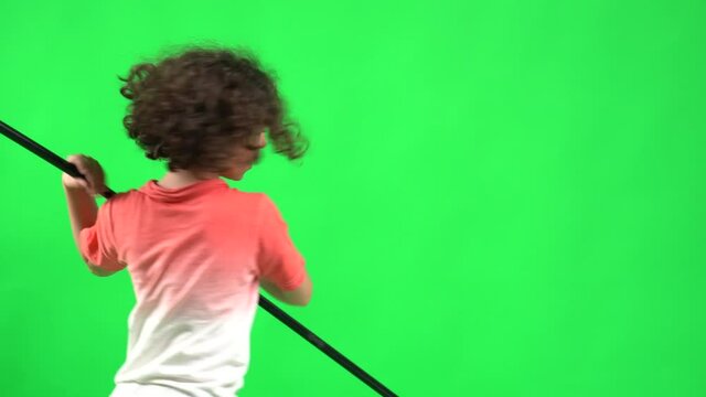 A cute kid with long brown curly hair, playing with a toy battle axe in front of the camera and behind a green screen.
