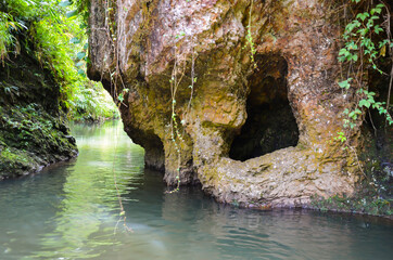 Beautiful scenery of lake water flowing through the hills and dark cave inside the mountain with shrub, green grasses and trees.