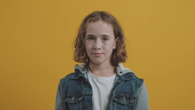 Isolated Handsome Caucasian School Boy Standing on a Yellow Coloured Background Looking Directly at the Camera, Smiling and Gesturing a Peace Sign With His Hand, Wearing a Cool Sweatshirt Jacket.
