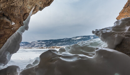 The granite rock is covered with bizarre ice layers. Through the exit of the cave you can see a frozen lake, a mountain range, a cloudy sky. Baikal