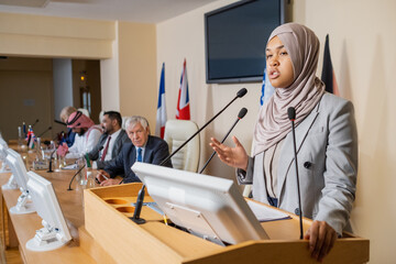 Confident young Muslim politician in hijab standing at rostrum and expressing her positiong while...