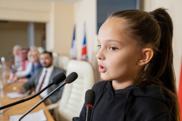 Little activist with ponytail speaking into microphone while addressing conference in front of...