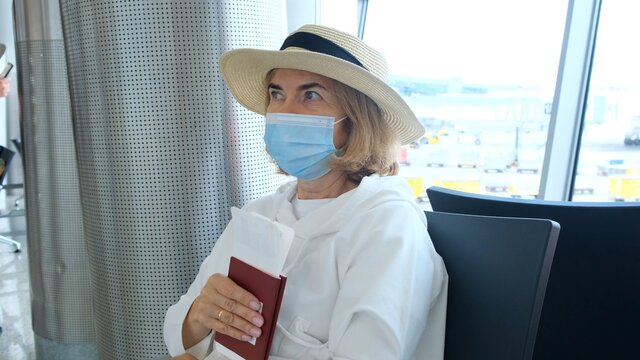 Adult Female Wearing A Hat And A Protective Mask Sits At The Check-in Counters In The Airport Terminal Awaiting The Departure Of The Flight Holding Air Tickets Due To Travel Restrictions Due To The