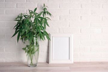 Cannabis plant in a vase with blank photo frame on a white brick wall. Copy space for text.