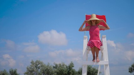 Lifeguard woman in a red dress sits on a white wooden rescue chair on the beach