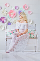 Beautiful pregnant young woman in a white dress sits on a bench. Smiling pregnant blonde in white interior.  Happy motherhood.