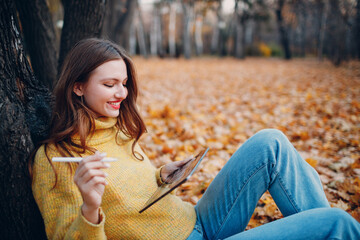 Young woman model sitting with tablet pc and painting with pencil stylus in autumn park with yellow foliage maple leaves.