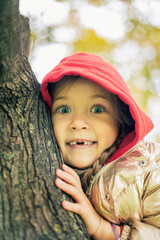 A girl in a gold jacket and red hood sits on a tree and smiles a toothless smile