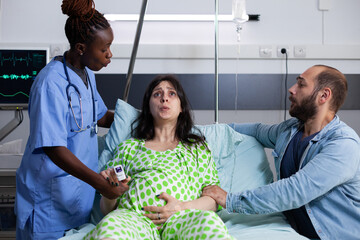 Pregnant woman in pain getting medical assistance from black nurse while sitting in hospital ward...
