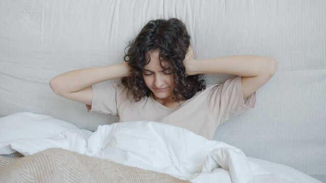 Unhappy young woman can't sleep because of loud noise covering ears with hands sitting in bed feeling angry and stressed. People and lifestyle concept.