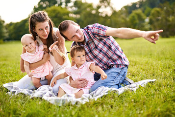 Fototapeta premium Happy family with young, millennial parents and two baby girls sitting on a picnic blanket and pointing on something in the distance to show it to their children - Outdoor lifestyle family concept