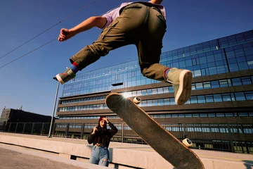 Foto auf Acrylglas A teenage boy performs tricks on a skateboard while a girl takes photos of him. © chika_milan