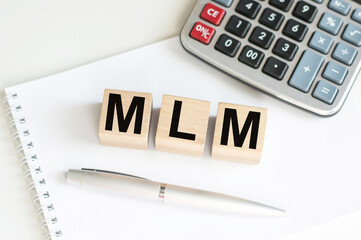 MLM Multi-Level Marketing written on a wooden cubes that lie on a white notebook next to a pen and a calculator