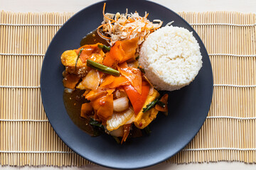 top view of portion of Com Dau Phu Vietnamese wok fried tofu with vegetables in sweet and sour sauce with boiled rice on black plate on wooden table