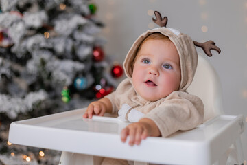 portrait of chubby cheeked baby in a jumpsuit with deer horns sits in a white high chair for feeding on the background of Christmas tree. winter New Year's concept. space for text. High quality photo