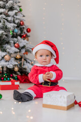 baby in a Santa costume is sitting next to the Christmas tree and playing with gift boxes. child opens a Christmas gift. winter new year's concept. space for text. High quality photo