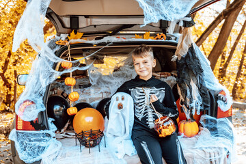 preteen caucasian boy in skeleton costume with bucket of sweets and cute poodle dog in ghost costume sits in trunk car decorated for Halloween with web, orange balloons and pumpkins, outdoor creative