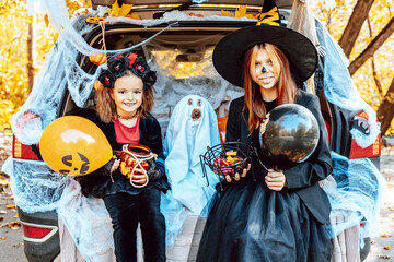 siblings teenage girl in witch costume and hat, cute little girl in spooky costume and cute poodle dog in ghost costume sits in trunk car decorated for Halloween with web, orange balloons and pumpkins