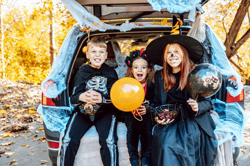 siblings boy in skeleton costume, teenage girl in witch costume and hat and cute little girl in spooky costume sits in trunk car decorated for Halloween with web, orange balloons and pumpkins, outdoor - obrazy, fototapety, plakaty