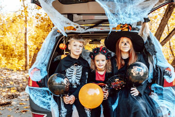 siblings boy in skeleton costume, teenage girl in witch costume and hat and cute little girl in spooky costume sits in trunk car decorated for Halloween with web, orange balloons and pumpkins, outdoor