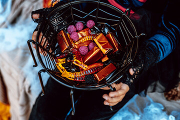 little girl in spooky costume and hat with bucket of sweets and cute poodle dog in ghost costume sits in trunk car decorated for Halloween with web, orange balloons and pumpkins, outdoor creative
