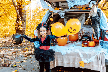 little girl in spooky cute costume and hat stands near trunk car decorated for Halloween with web, orange balloons and pumpkins, outdoor creative activity concept in autumn - obrazy, fototapety, plakaty