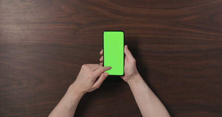 Top view man hands hold smartphone with green screen over black walnut wood table