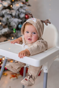 chubby cheeked baby in a jumpsuit with deer horns sits in a white high chair for feeding on the background of a Christmas tree. winter New Year's concept. space for text. High quality photo